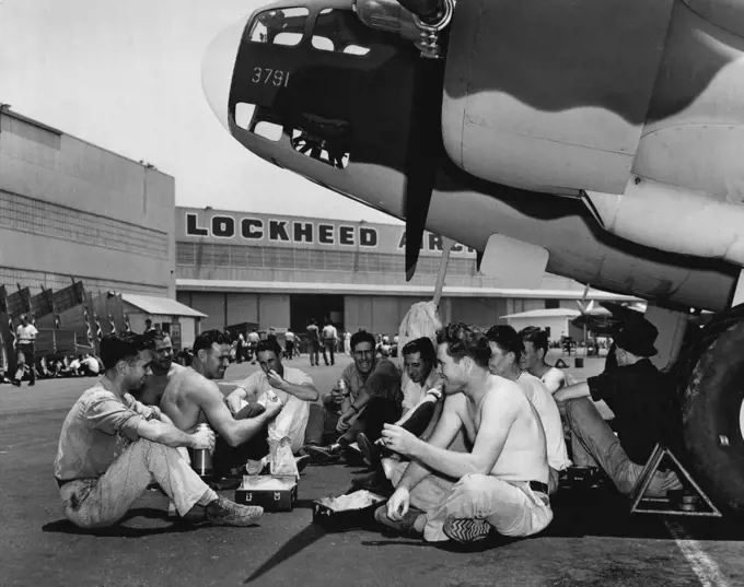 A bunch of the boys eat an Al Fresco lunch under the nose of a Lockheed Bomber they helped to build. They're kidding Palmer, (third from left) about his build. "Hey Superman!" one of them chortles between bites," when are you Gonna fly one o' these bombers to England.... on your back, I mean". July 18, 1941. (Photo by ACME).