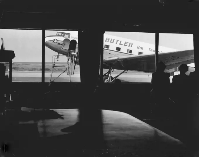 Passengers view a DC3 of ***** BAT fleet through the windows of the terminal at Mascot. November 30, 1955.