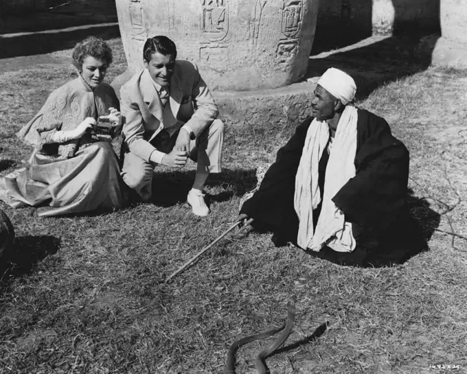 Egyptian Scenes... Between scenes of M-G-M's "Valley of the Kings" in Egypt, Eleanor Parker and Carlos Thompson look on as a local snake charmer in Luxor puts his pet cobra through his daily exercises. Robert Taylor co-stars in the exciting adventure film, most of which was made in Egypt. Robert Pirosh directed. July 3, 1954.