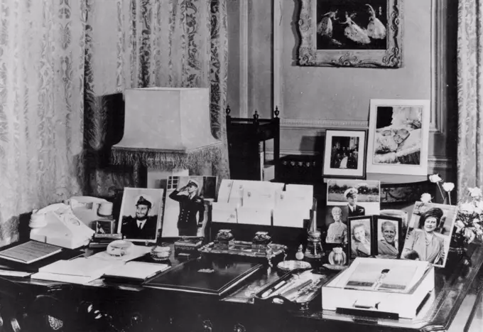 Clarence House - Princess Elizabeth's London Home -- Princess Elizabeth's writing desk in her Sitting Room at Clarence House, the London residence of the Princess and the Duke of Edinburgh. The desk is of the Chippendale period. The photographs are of the Queen, Queen Mary, Princess Margaret and several of the Duke of Edinburgh, including one taken during the war when he wore a beard. Beyond are photographs taken at the christening of Prince Charles, and a painting of the ballet Le Iac des Cygnes, by Oliver Messel. October 28, 1949.