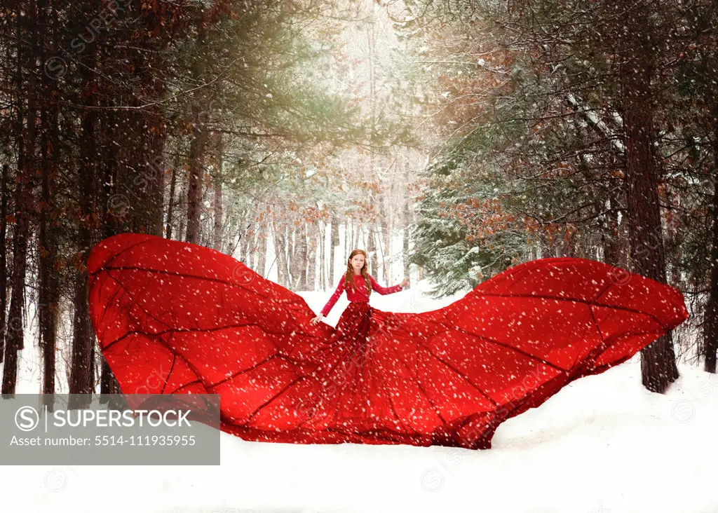 Young redhead girl in flowing red dress in forest with snowfall.