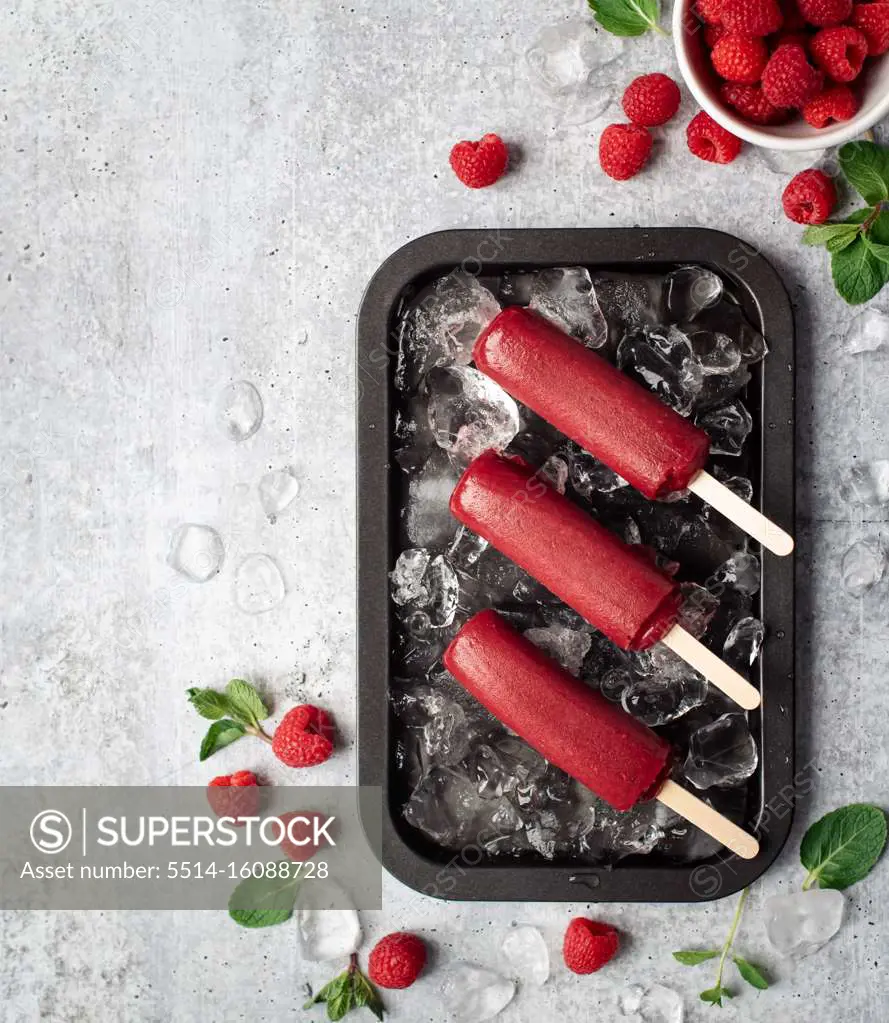 Raspberry popsicles on a tray with ice and fruit on grey background.