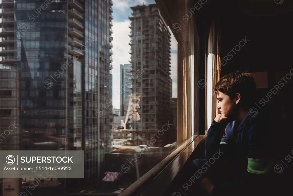 Tween boy looking out a window at tall buildings of the city outside.