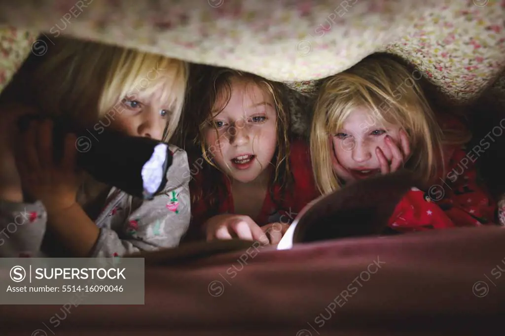 Three little girls reading book under blanket with flashlight