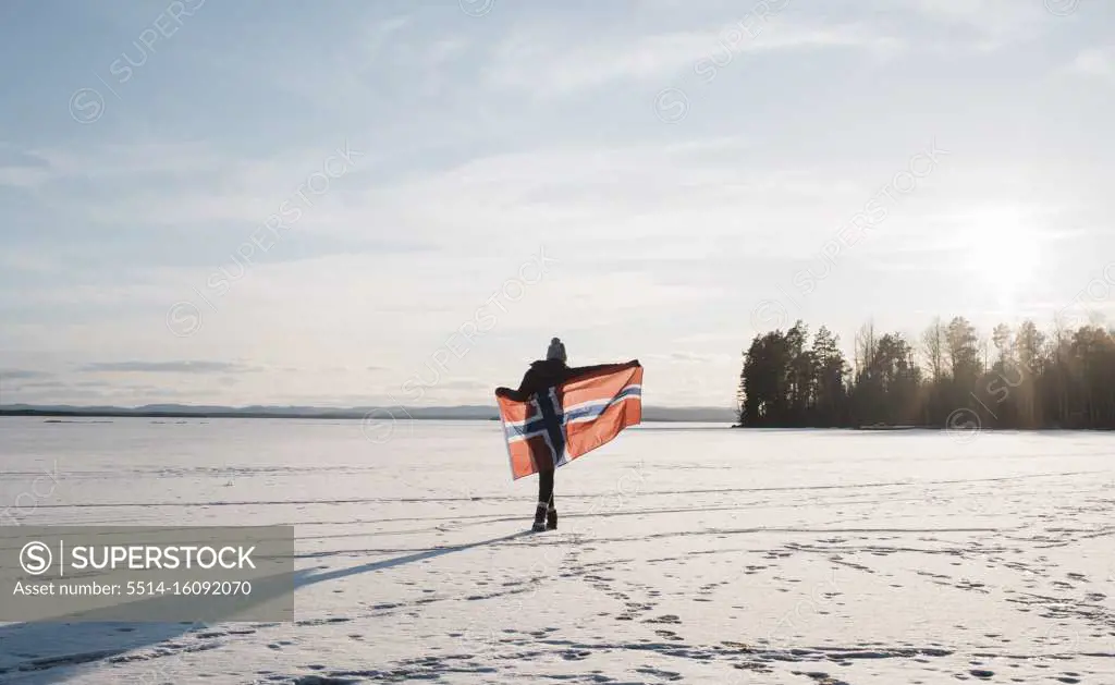 woman walking across a frozen lake holding a Norwegian flag at sunset