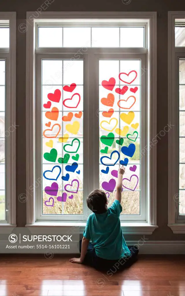 Boy sitting on floor by a window covered in a rainbow of paper hearts.