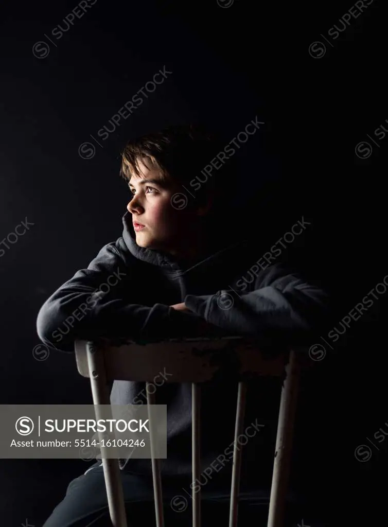 Low key portrait of adolescent boy sitting on a chair in a dark room.