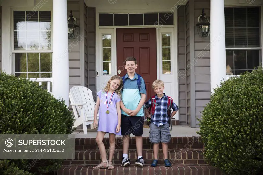 Three Smiling Happy Siblings With Backpacks Stand on Brick Front Steps