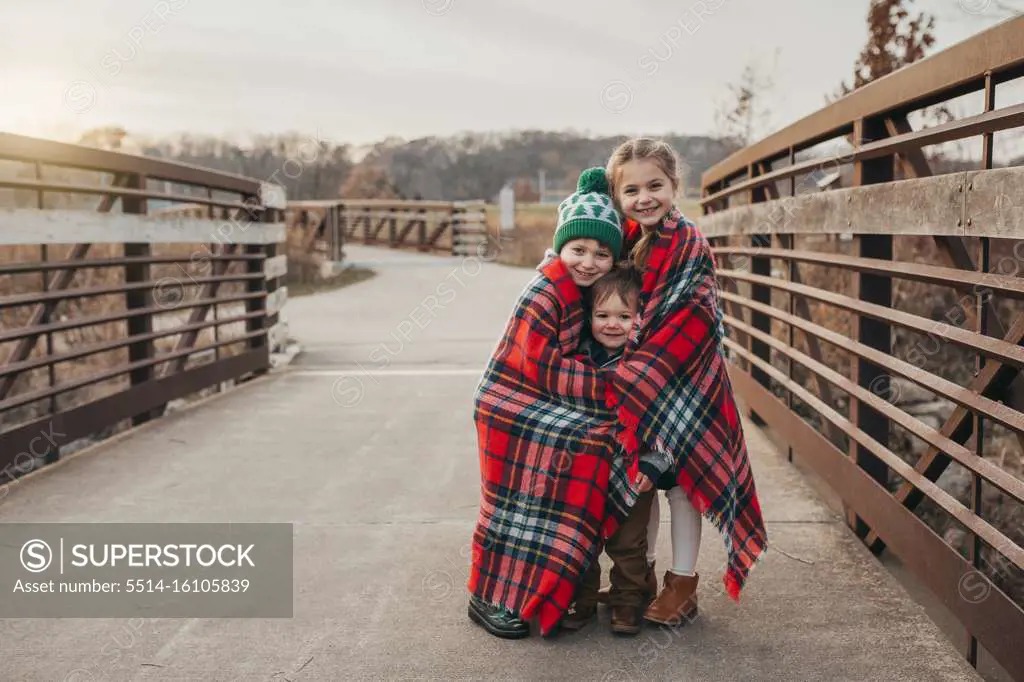 Siblings wrapped in Christmas plaid blanket on bridge at sunset