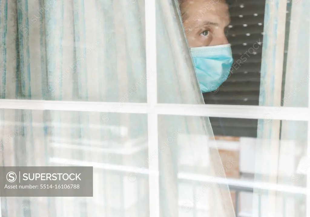 Photo of a Middle-aged white man with medical face mask looking out of the window during coronavirus quarantine