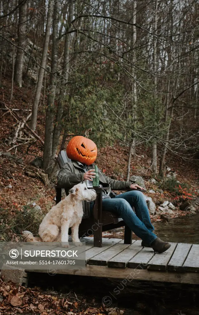 Man wearing scary pumpkin head for Halloween sitting on dock with dog.