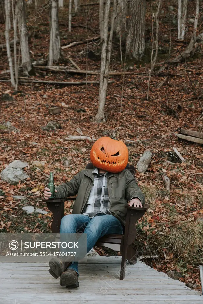 Man wearing scary pumpkin head at Halloween sitting on dock with beer.