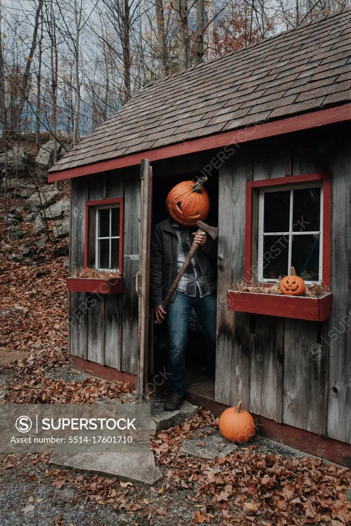 Man wearing scary carved pumpkin head in shed with axe for Halloween.