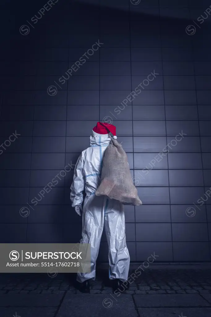 Back View of a volunteer, dressed in the coverall used against coronavirus, the face mask, and the beard and hat of Santa Claus, standing in front of the wall of a hospital, ready to enter to