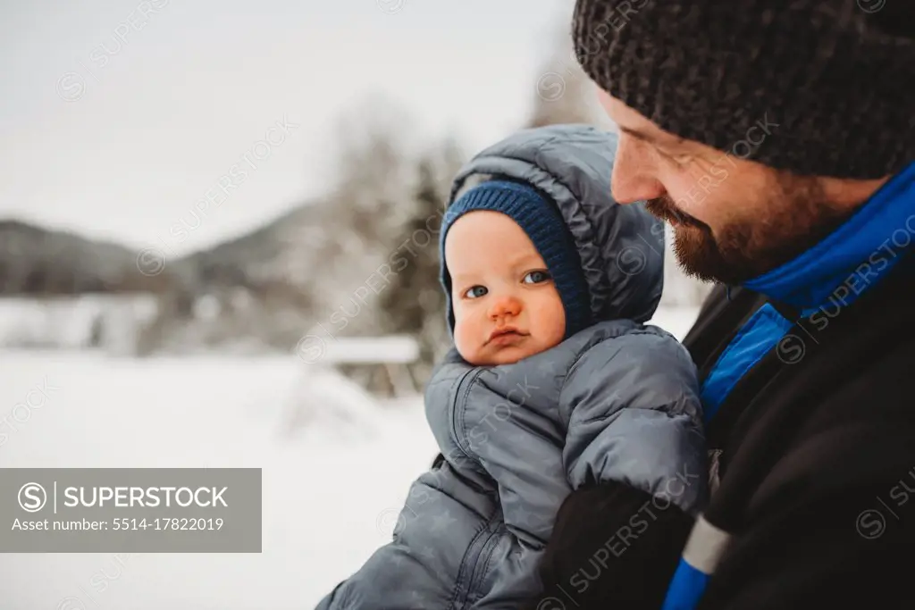 close up of Dad holding baby wearing snowsuit in winter with snow