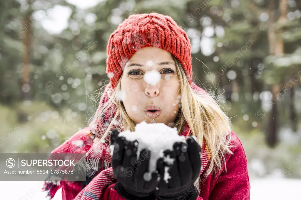 Portrait of happy young woman playing with snow in winter, blowing snowflakes to camera, copy space