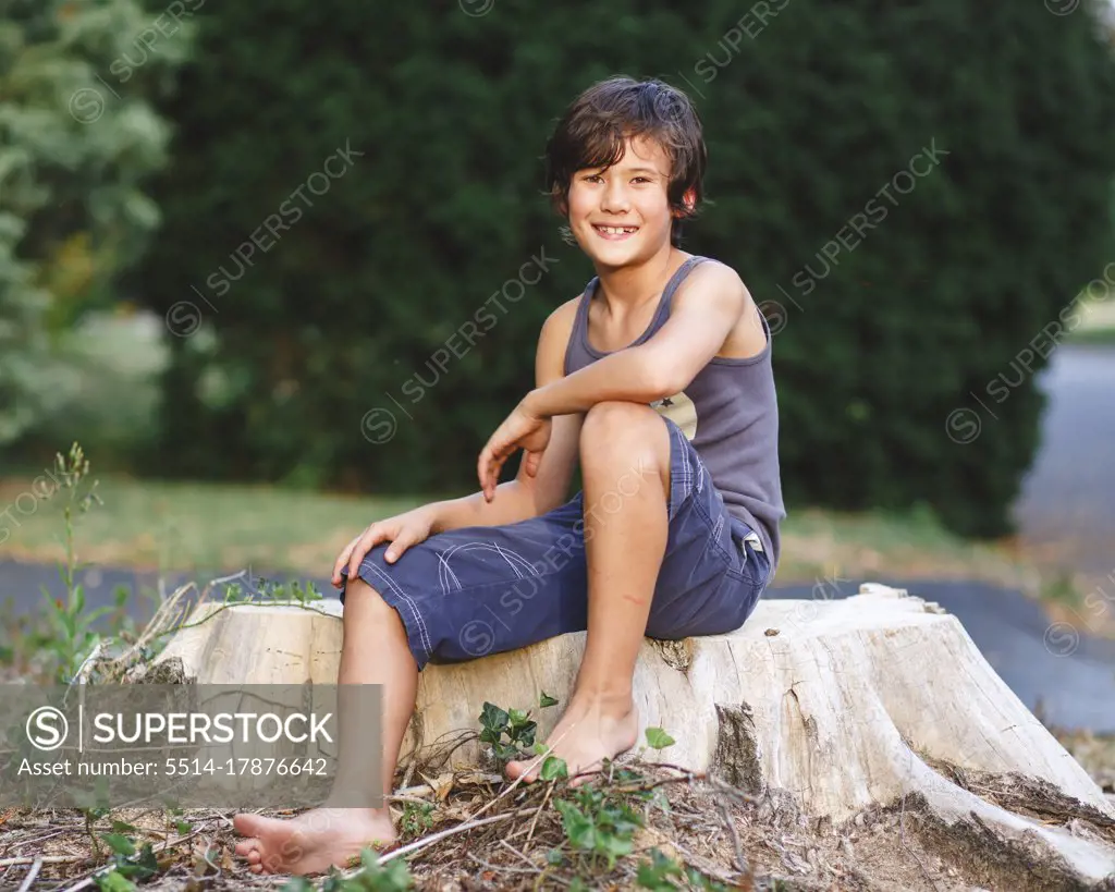 portrait of happy barefoot boy with golden skin sitting on tree stump