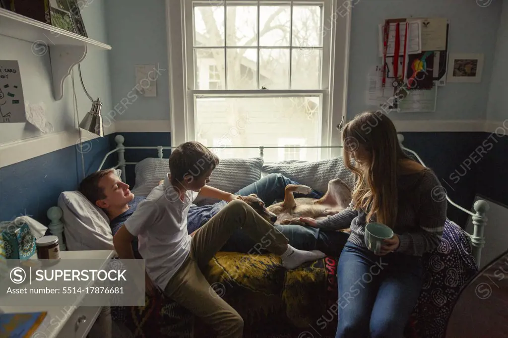 three siblings sit on a bed cuddling with a cute family dog in bedroom