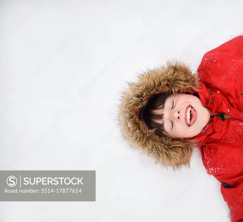 Overhead of happy boy in red coat with furry hood laying in the snow.