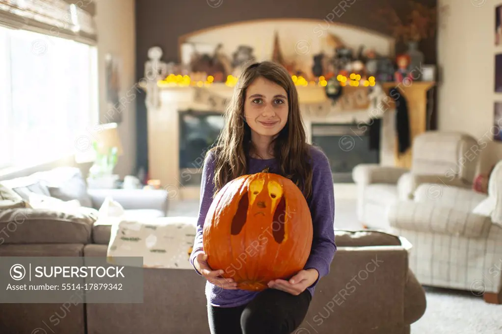 Tween girl 10-12 years old holds up carved pumpkin in living room