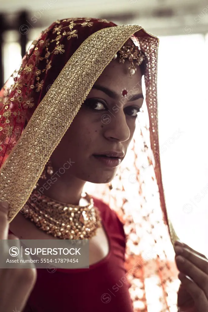 Young Indian woman pulls on red wedding veil in window light