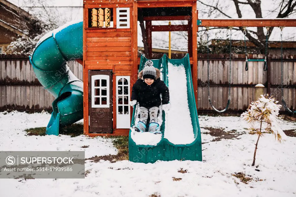 Young boy sliding on snow covered swingset in backyard at home