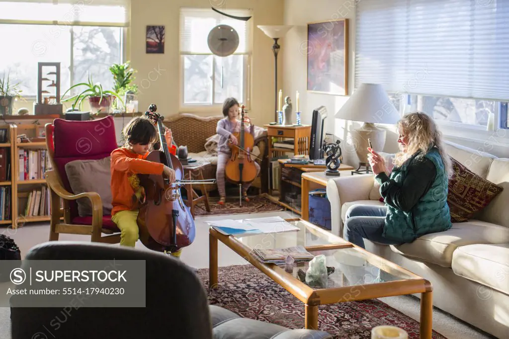 Two children perform cello at home and grandmother records with phone