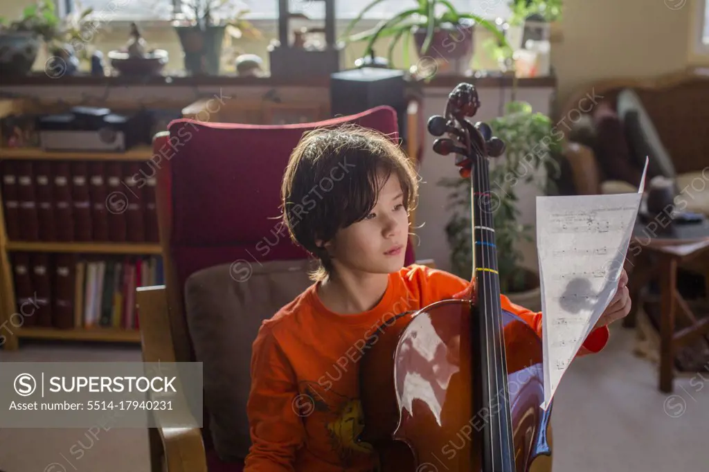 A boy sits in beautiful light with a cello reading sheet music