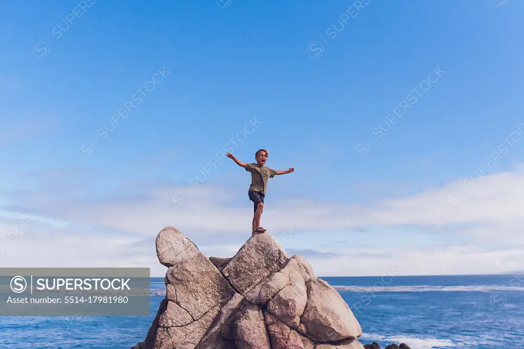 Boy screaming on top of a rocky mountain by the ocean.