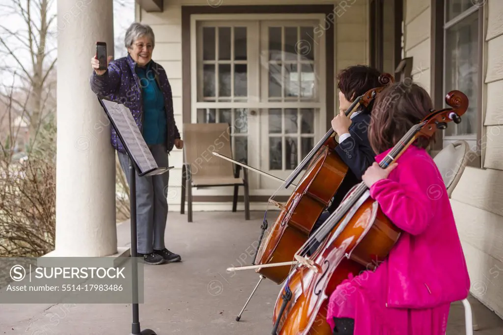 An elderly woman records two children giving cello concert on porch