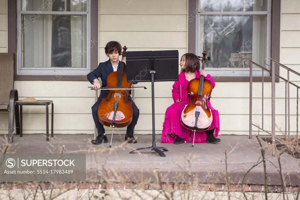 A tween boy sits on porch playing cello in suit while sister watches