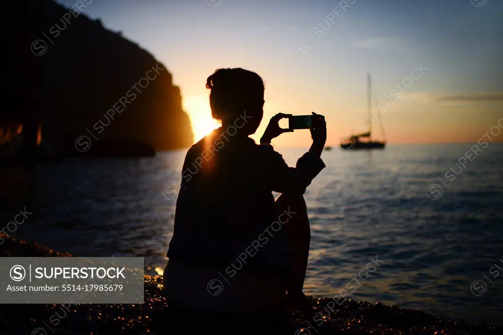 Silhouette of a young woman using a smartphone to capture sunset