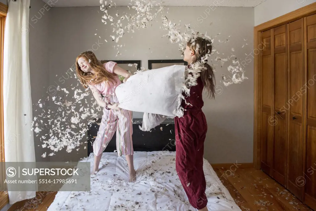 Two happy young girls having a feather pillow fight on the bed.