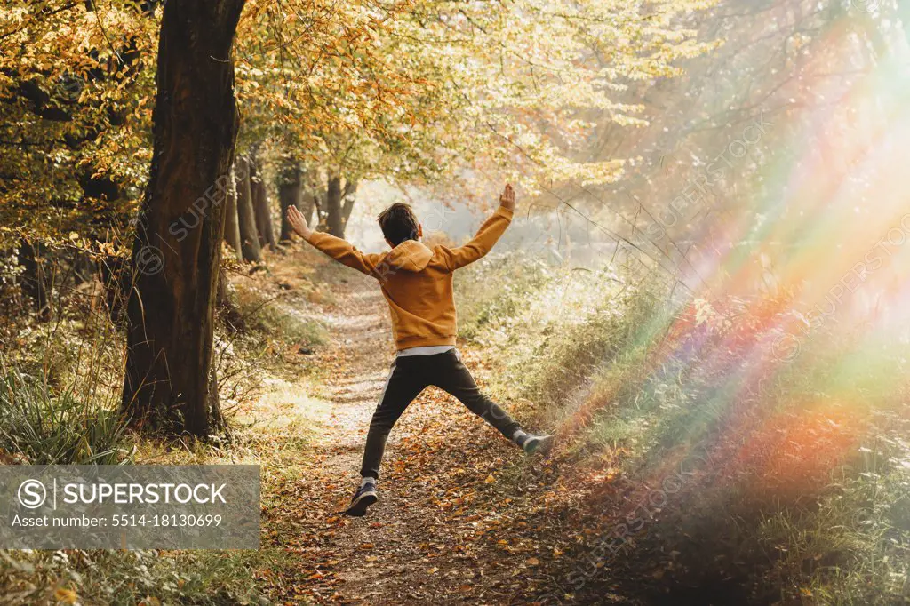 Boy leaping in the air on path under tree with rainbow light flare