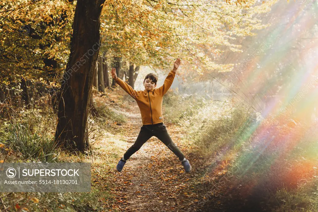 Boy leaping in the air on path under tree with rainbow light flare