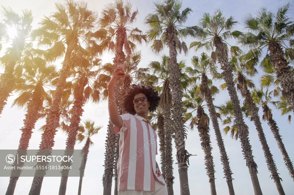 Latino in his 20s taking a selfie among the palm trees on the beach. Dark-skinned man taking a selfie of himself while standing.