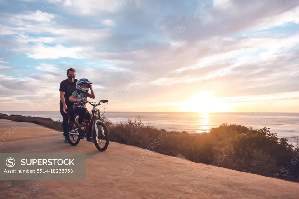 Father helping son ride his bike on a coastal trail at sunset.