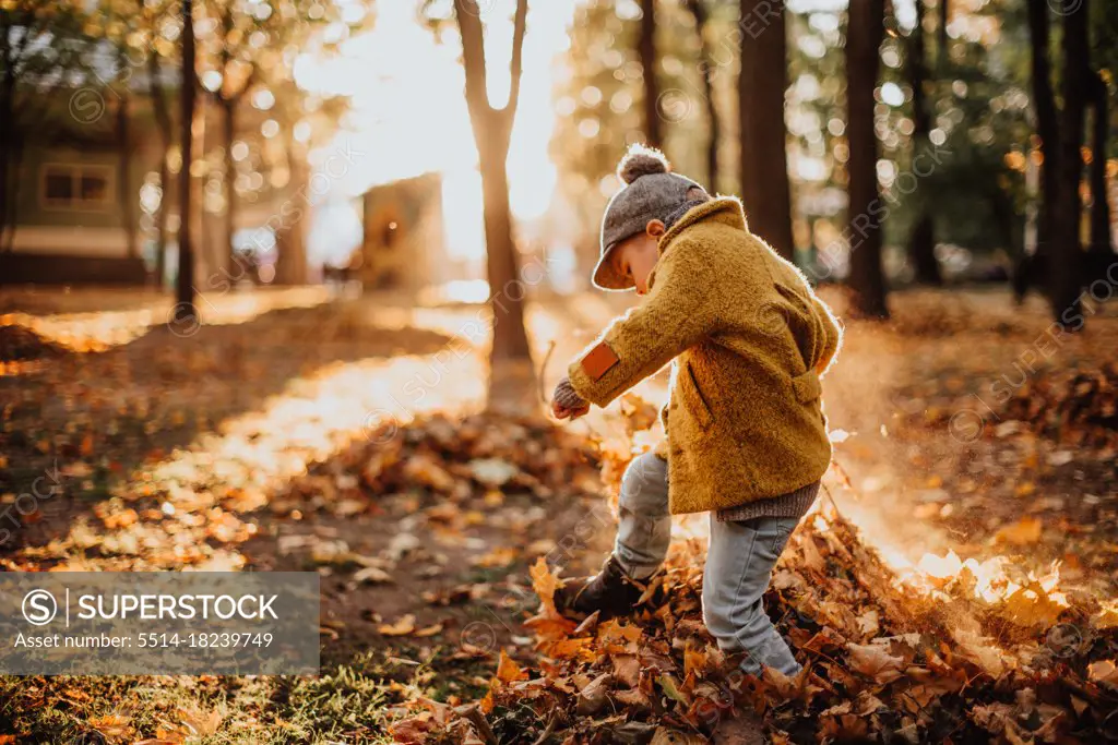 Stylish boy having fun in autumn city park. Happy kid walking among fallen leaves. Kids fashion. Boy wearing trendy yellow coat, cap and scarf. Smiling young boy outdoors. Kid jumping and run