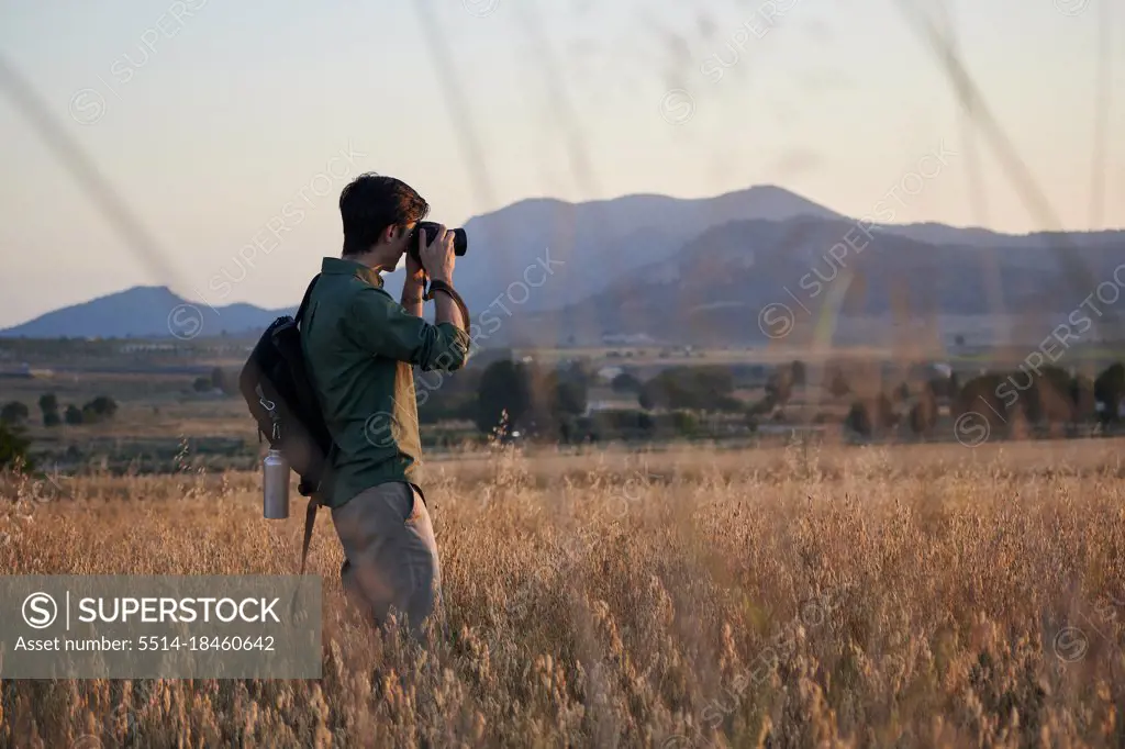 Man photographer with a backpack takes a photo in a meadow at sunset