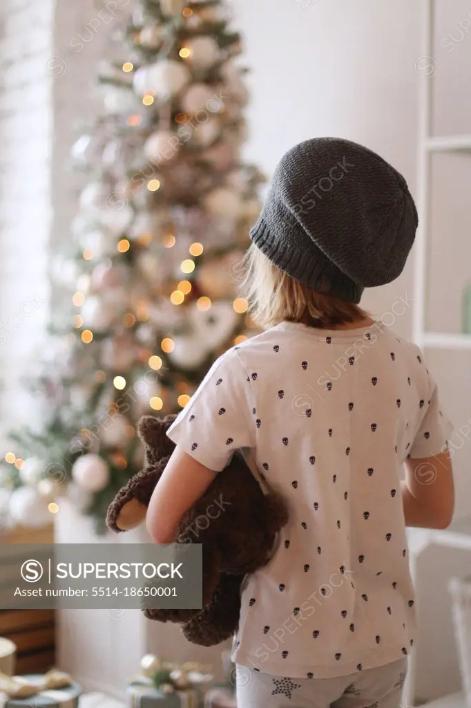 a child in a hat stands in front of a christmas tree