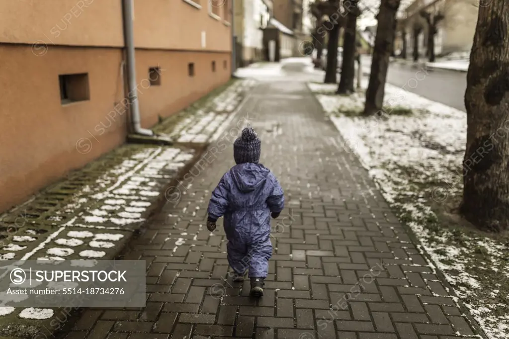 Little boy in blue onesie and winter hat walking on pavement