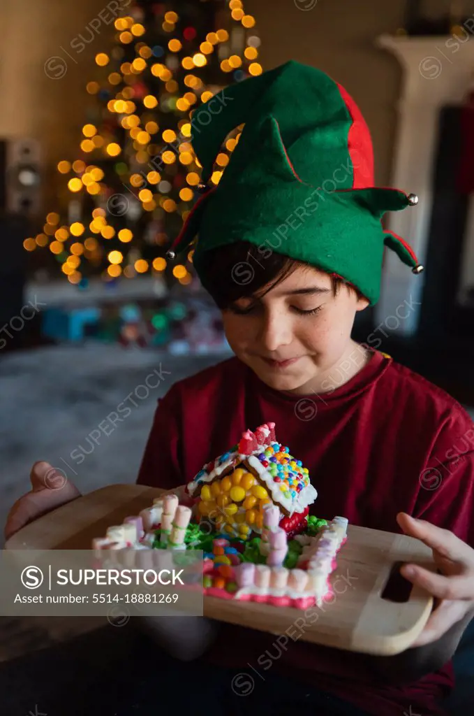 Happy boy in elf hat looking at gingerbread house at Christmastime.