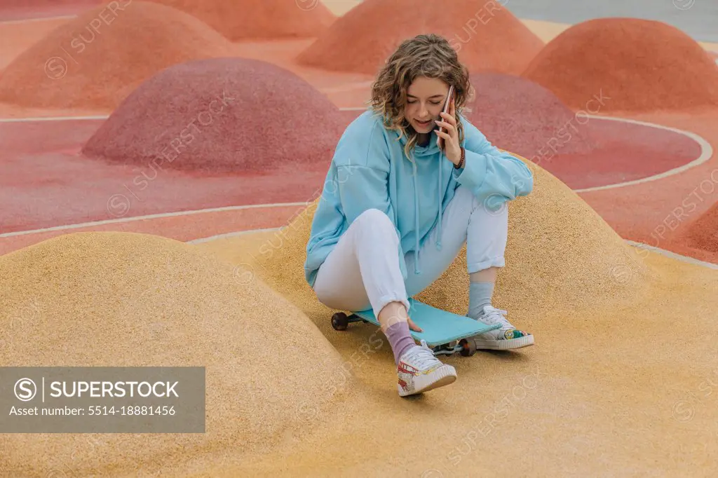 Young hipster millennial woman talking on phone sit on skateboard.