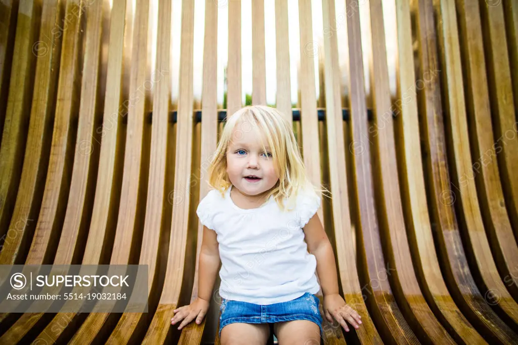 Cute blonde girl sitting on wooden bench at park.