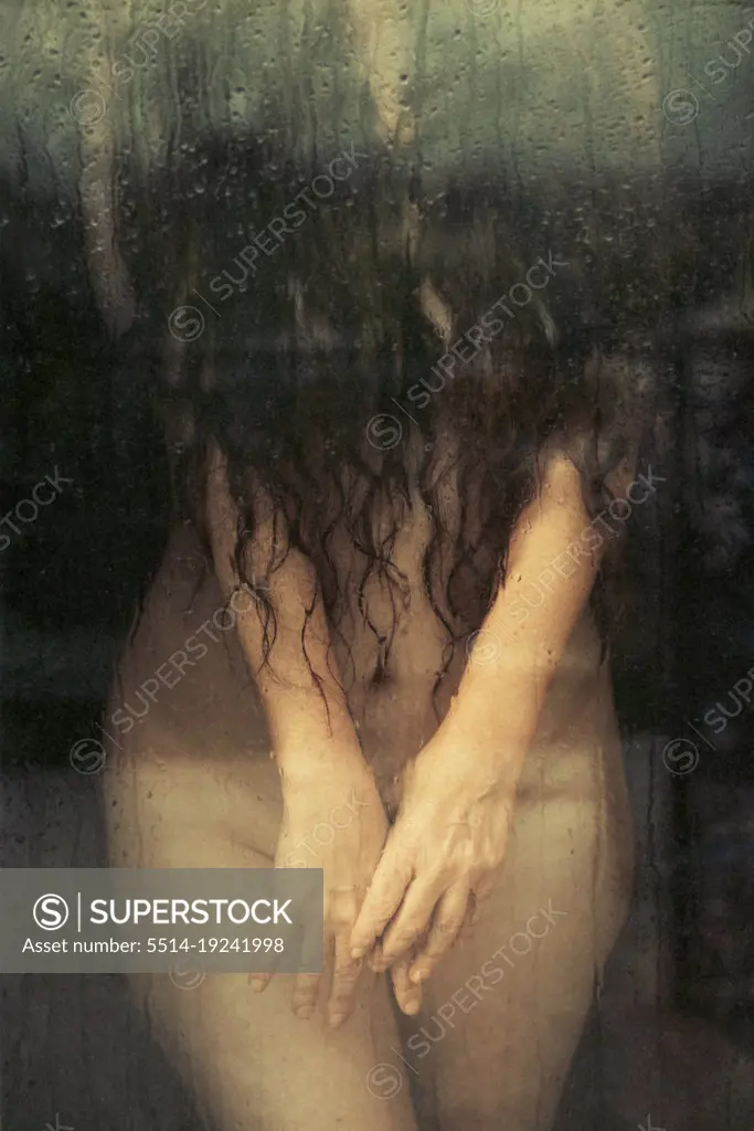 a woman standing behind glass covering her naked body with her hands