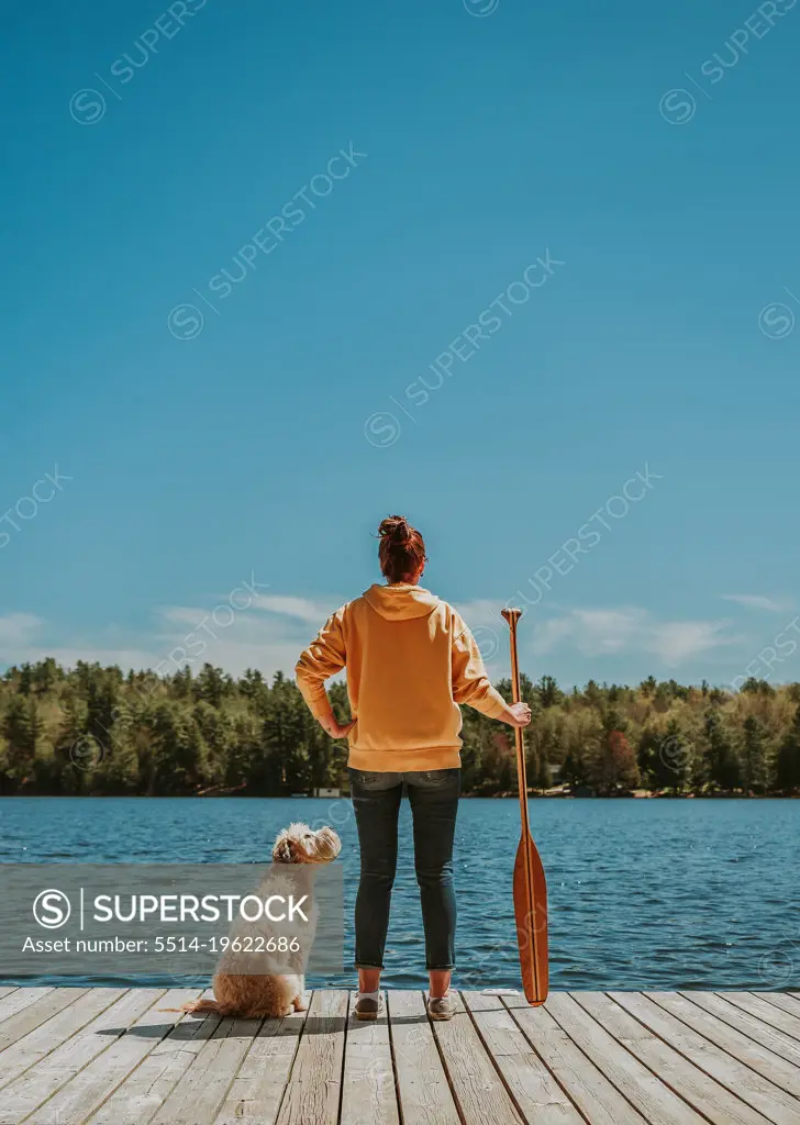 Woman standing on dock with dog holding paddle looking out at lake.