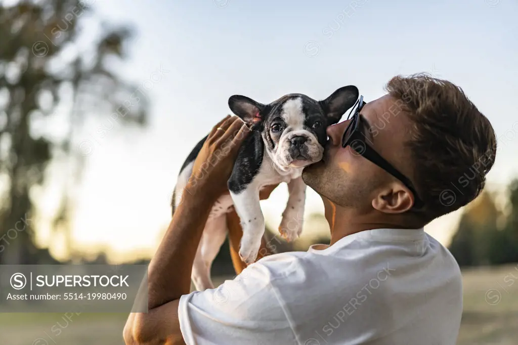 Handsome young man holding up and kissing a french bulldog.