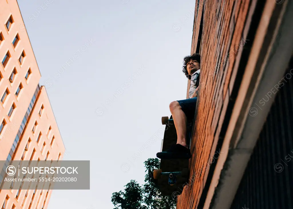 man sat in the window of a building with his skateboard at sunset