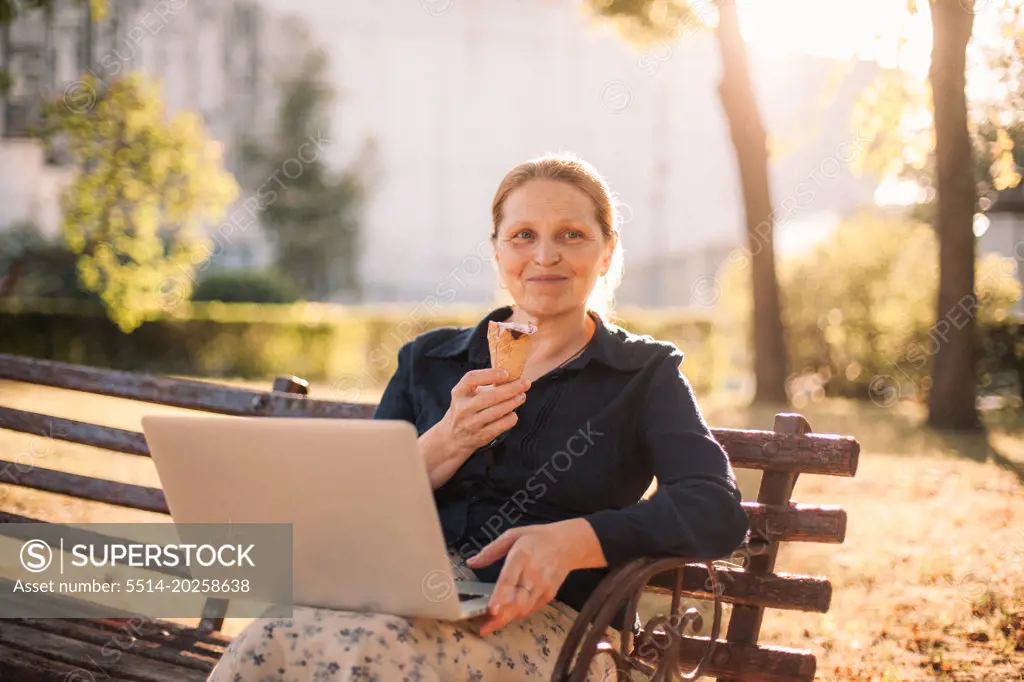 Happy senior business woman holding laptop and ice cream in park