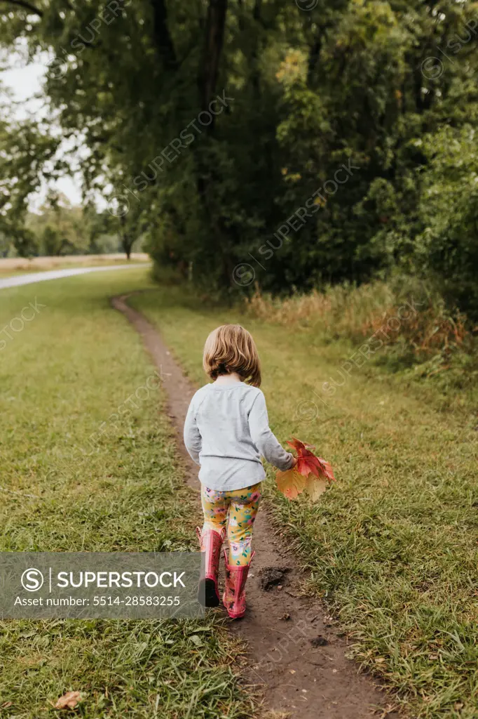 Young girl walks on dirt trail while holding fall leaves in hand